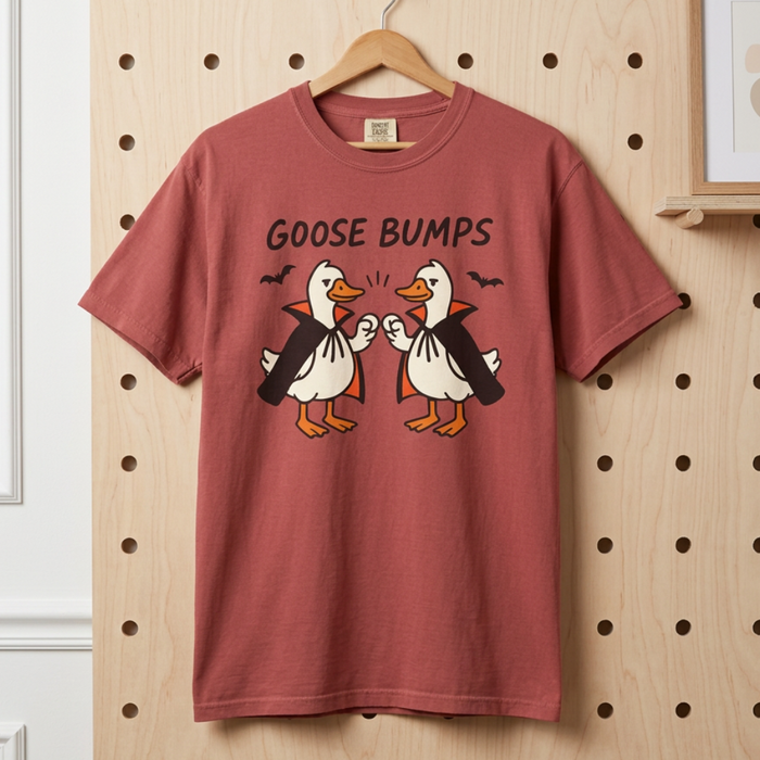 A dusty rose short-sleeve t-shirt hanging on a wooden hanger against a light wood pegboard wall. The shirt features a graphic of two cartoon geese bumping fists, with the text 'GOOSE BUMPS' above them.