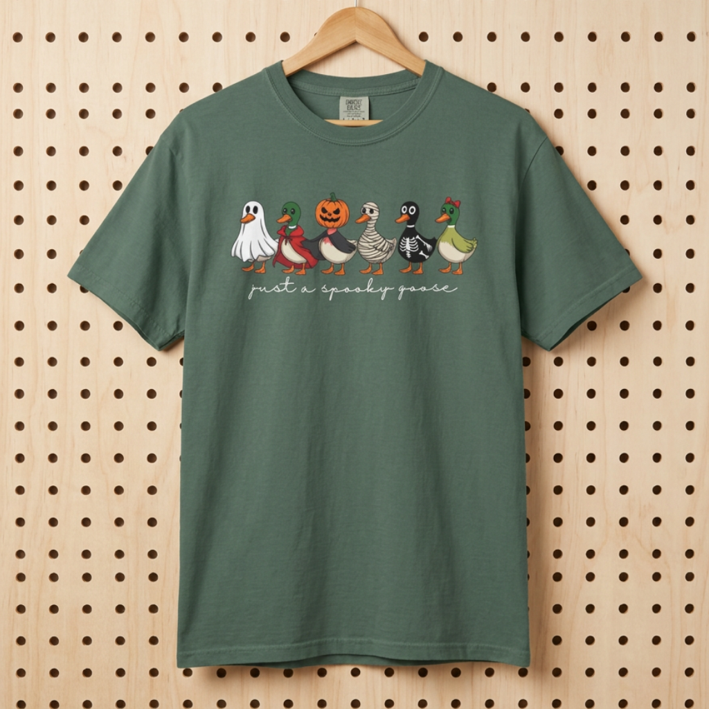A blue spruce short-sleeve t-shirt hanging on a wooden hanger against a light wood pegboard. The shirt features a graphic of 6 cartoon ducks dressed in Halloween costumes (ghost, pumpkin, acorn, witch, spider, zombie, and turtle) with the text 'just a spooky goose' printed in white script underneath