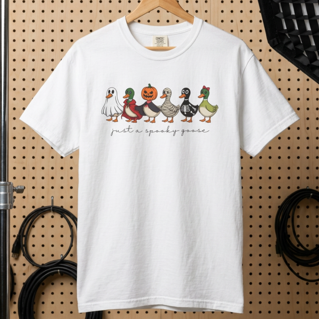 A white short-sleeve graphic t-shirt hanging on a wooden hanger against a tan pegboard backdrop. The shirt features a row of cartoon ducks in Halloween costumes with the text 'just a spooky game' printed below them.