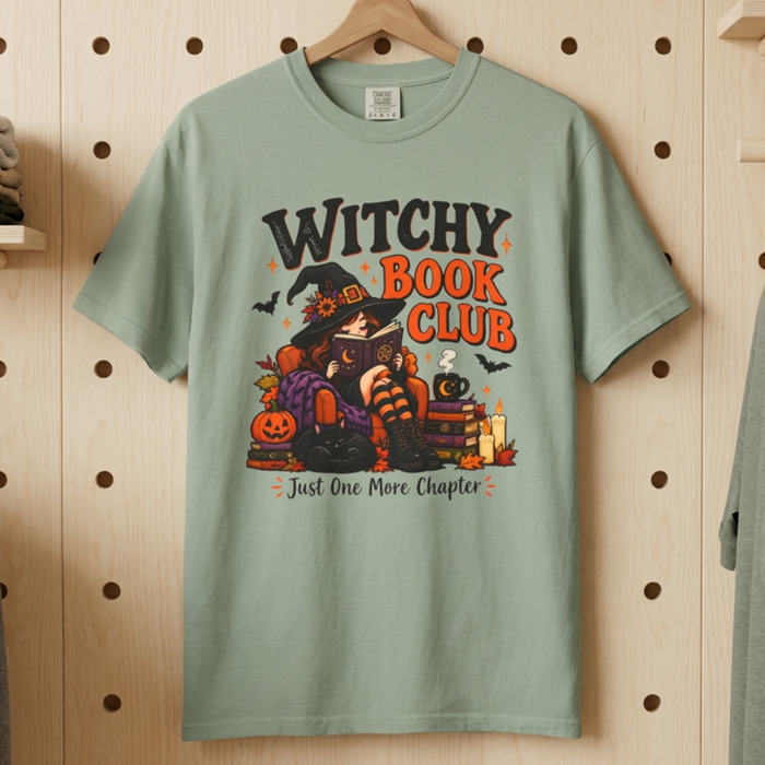 A bay graphic t-shirt hanging on a wooden hanger against a light wood pegboard wall. The shirt features a colorful graphic of a witch sitting on a pumpkin tree with the text 'WITCHY BOOK CLUB' and 'Just One More Chapter'