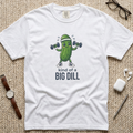 A white graphic t-shirt. The t-shirt features a cartoon pickle character lifting dumbbells with the text 'kind of a BIG DILL' printed below.