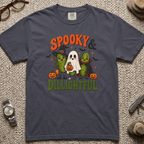 A dark slate blue graphic t-shirt laid flat on a textured beige carpet, surrounded by lifestyle props including a pair of eyeglasses, a watch, a small hat, and a pocket knife. The t-shirt features a Halloween graphic with the text 'SPOOKY & DILIGHTFUL' in orange and green, depicting two cacti dressed as ghosts with pumpkins.