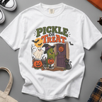 A white short-sleeve t-shirt with a colorful Halloween graphic reading 'PICKLE OR TREAT' and featuring a green pickle character, a ghost, pumpkins, and a door, laid flat on a beige fabric background.