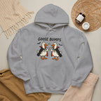 A light gray pullover hoodie laid flat with a graphic of two cartoon geese in vampire outfits and the text 'GOOSE BUMPS' above them. The hoodie is styled in a flat lay with a beige knit blanket, tan sweater, beige pants, and a woven placemat with small jars on a light neutral background.