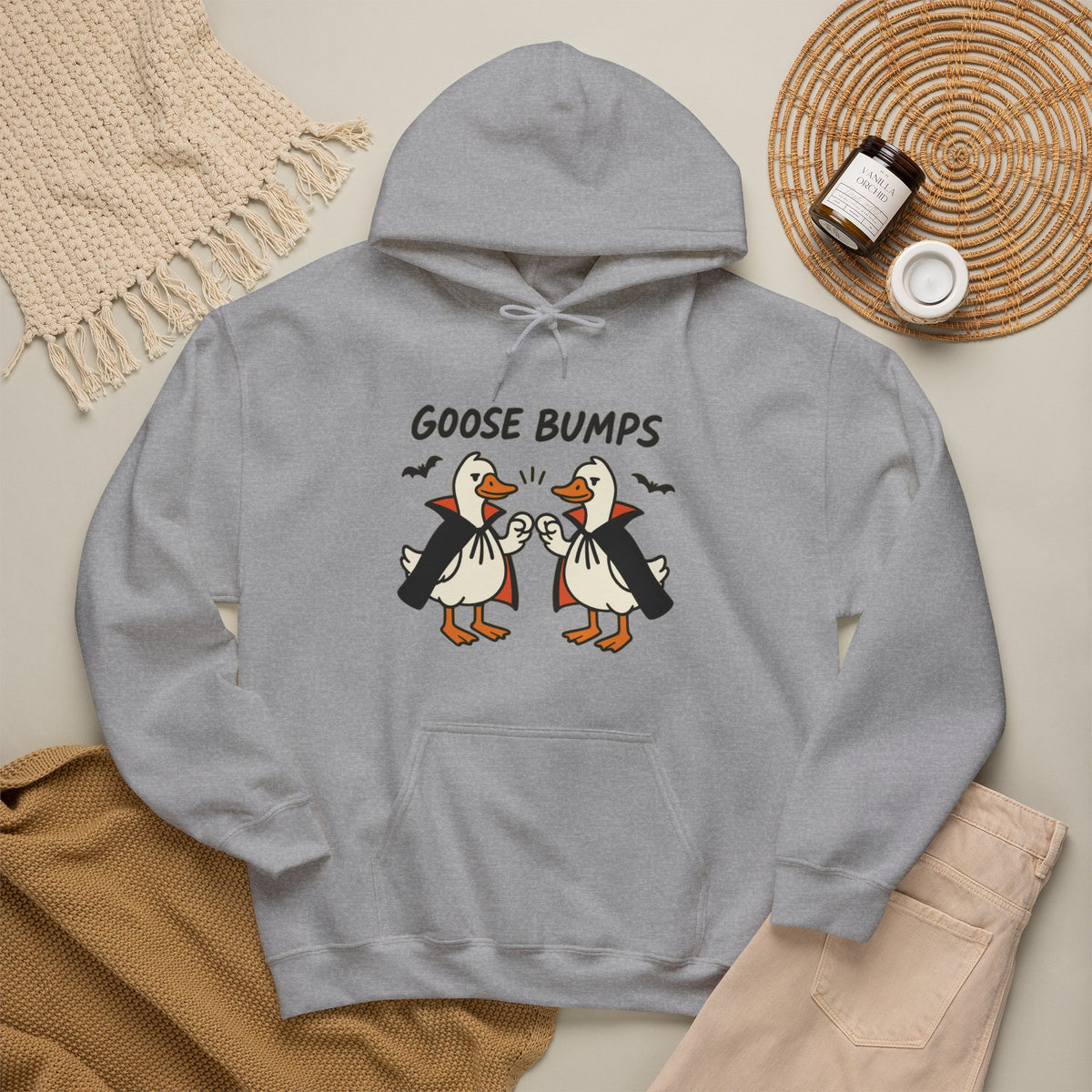 A light gray pullover hoodie laid flat with a graphic of two cartoon geese in vampire outfits and the text 'GOOSE BUMPS' above them. The hoodie is styled in a flat lay with a beige knit blanket, tan sweater, beige pants, and a woven placemat with small jars on a light neutral background.