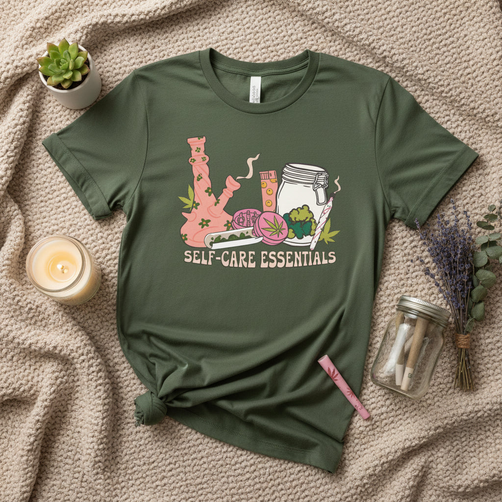 An olive green short-sleeve t-shirt with a colorful graphic on the front showing a pink bong, a jar, a pill bottle, and a green leaf, with the text 'SELF-CARE ESSENTIALS' printed below.