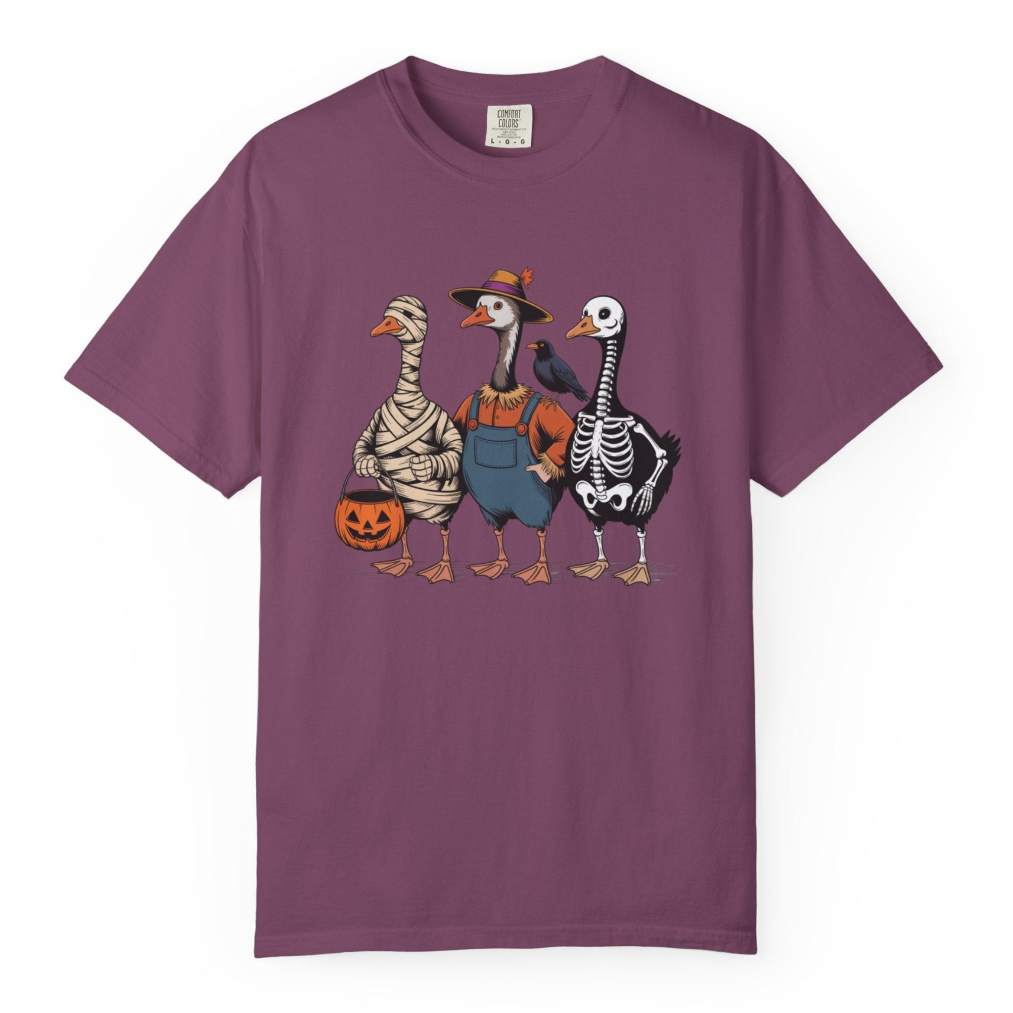 A purple short-sleeve crewneck t-shirt featuring a graphic print of three geese dressed in Halloween costumes (as mummy, skeleton, and in blue overalls), set against a plain white background.