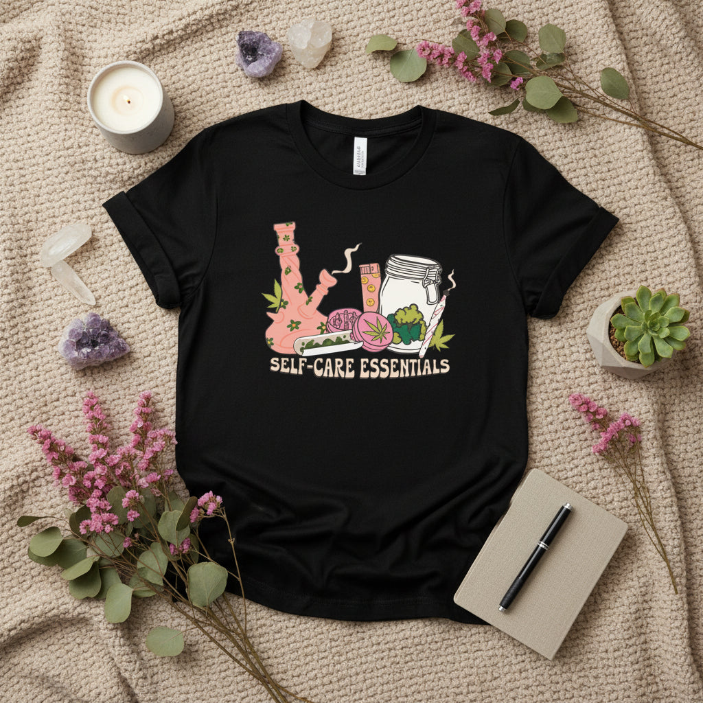 A black short-sleeve t-shirt with a colorful graphic on the front showing self-care items like a pink bong, a pill bottle, a jar, and a green leaf, with the text 'SELF-CARE ESSENTIALS' underneath.