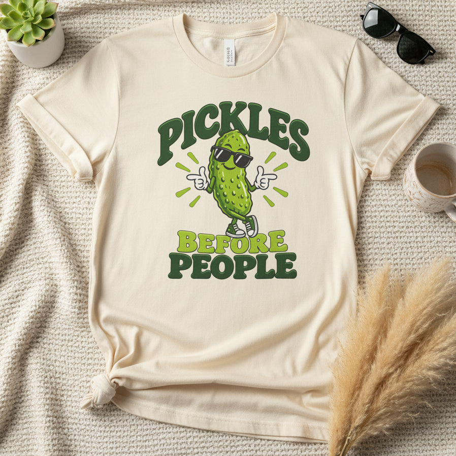 An off-white short-sleeve t-shirt with a graphic print of a cartoon pickle wearing sunglasses and pointing, with the text 'PICKLES BEFORE PEOPLE' in dark green and lime green.