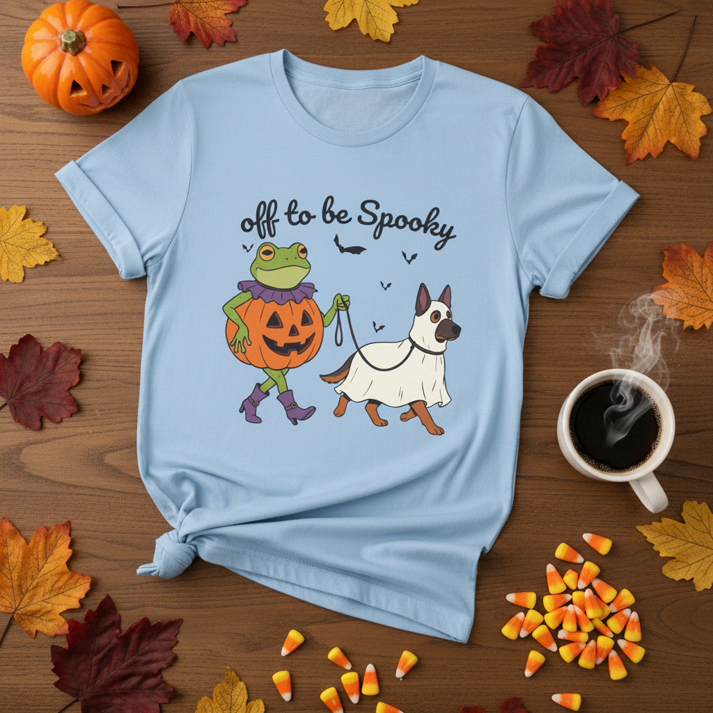 A light blue short-sleeve crewneck t-shirt featuring a cartoon graphic of a green frog in a pumpkin costume and a white dog in a ghost costume walking together, with the text 'off to be Spooky' above them.