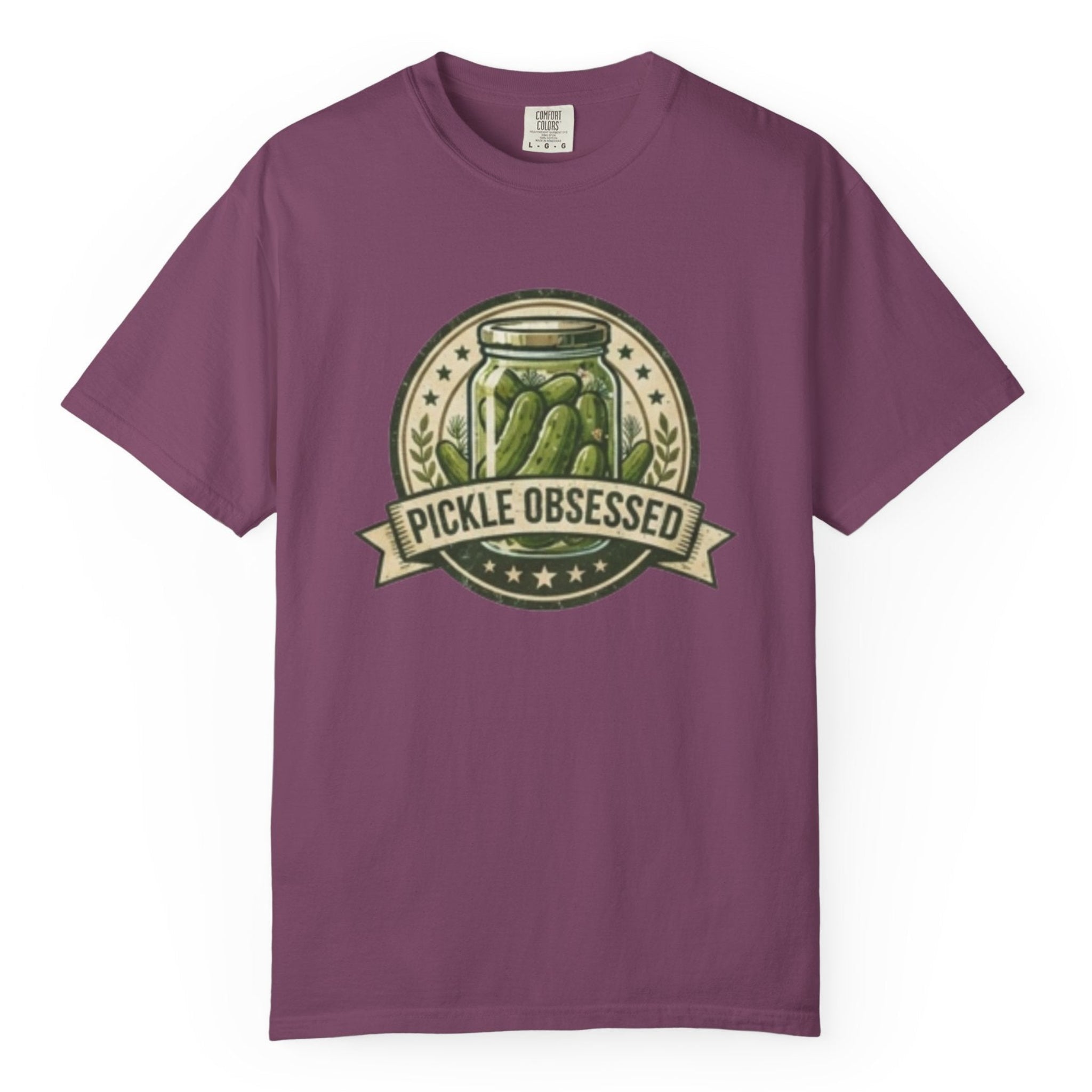 A purple short-sleeve t-shirt hanging on a wooden hanger against a light wood pegboard wall. The shirt features a circular graphic print with a jar of pickled green onions and the words 'PICKLE OBSESSED' in a banner.