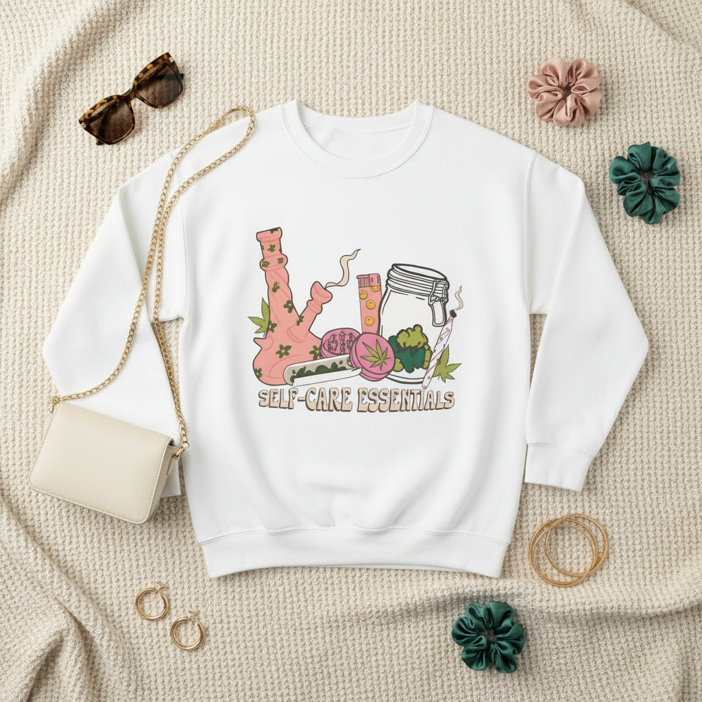 A white crewneck sweatshirt featuring a colorful graphic of a pink bong, a glass jar, a skull, and a flower with the text 'SELF-CARE ESSENTIALS' printed below, displayed on a clean white background.