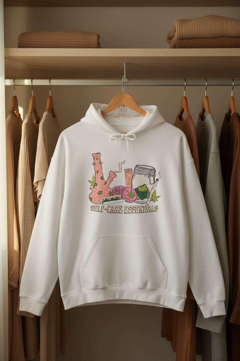 A white pullover hoodie with a colorful graphic on the chest depicting a pink bong, a pink cup, a glass jar, and green herbs, with the text 'SELF-CARE ESSENTIALS' printed below.