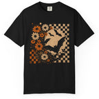 A black short-sleeve crewneck t-shirt featuring a distressed graphic print of a witch's hat, bats, orange and white flowers, and a checkered pattern on the front, displayed against a solid white background.