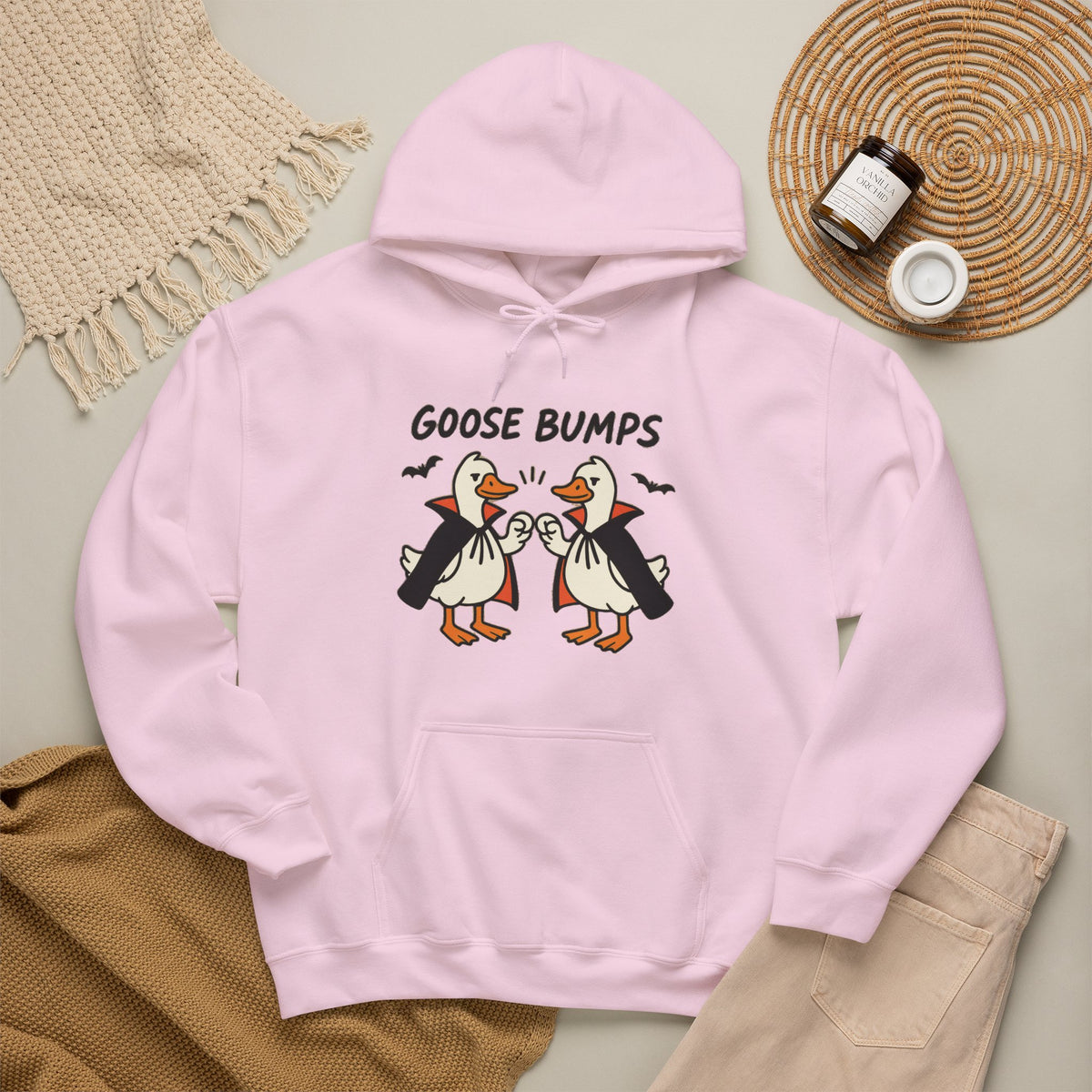 A light pink hoodie with a graphic of two geese in black and white costumes with orange beaks, under the text 'GOOSE BUMPS'. The hoodie is styled in a flat-lay arrangement with a beige knit blanket, a brown knit sweater, light pink pants, and a small jar on a woven mat.