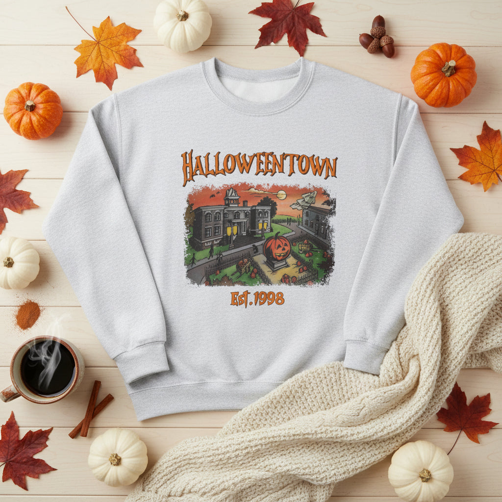 A light heather gray crewneck sweatshirt featuring a Halloween-themed graphic print on the front with the text 'HALLOWEENTOWN' and 'Est. 1998' surrounding an illustration of haunted houses and a jack-o'-lantern.