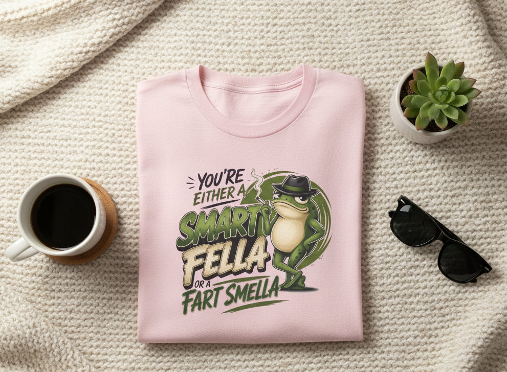 A light pink short-sleeve crewneck t-shirt featuring a graphic print of a frog wearing a hat and the text 'YOU'RE EITHER A SMART FELLA OR FART SMELLA'.