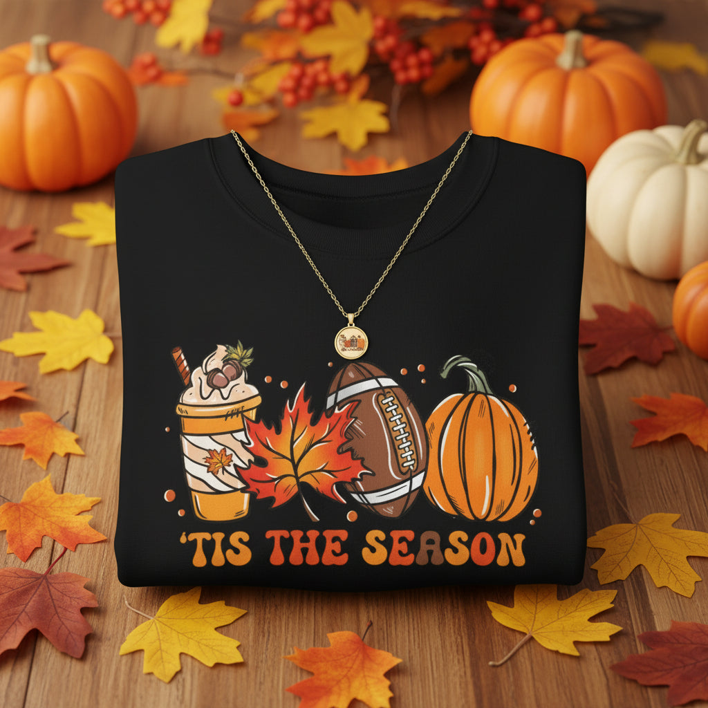 A black crewneck sweatshirt featuring a festive autumn graphic with a pumpkin, football, hot chocolate cup, maple leaf, and the text 'TIS THE SEASON' 