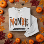 A light heather gray crewneck sweatshirt featuring a Halloween-themed graphic on the chest that reads 'MOMBIE' in dripping orange, black, and leopard-print lettering, with small bats and stars.