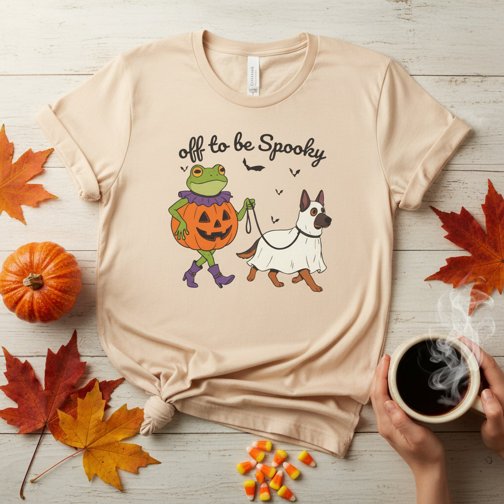 A soft cream crewneck t-shirt featuring a Halloween graphic of a green frog in a pumpkin costume walking a white dog dressed in a ghost costume, with the text 'off to be Spooky' above them.