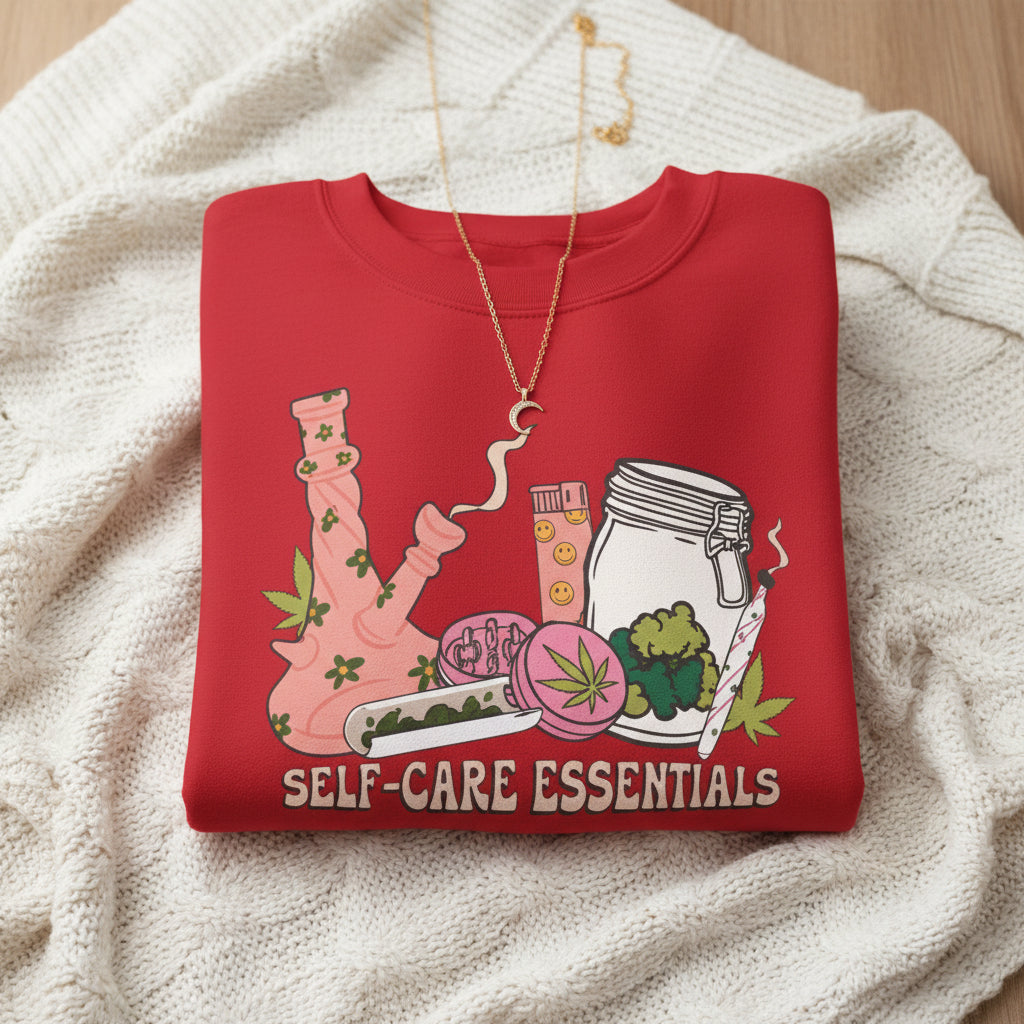 A red crewneck sweatshirt featuring a colorful graphic of self-care and healing items, including a smoking water pipe, a jar, a slice of watermelon, and a cup, with the text 'SELF-CARE ESSENTIALS' printed below.