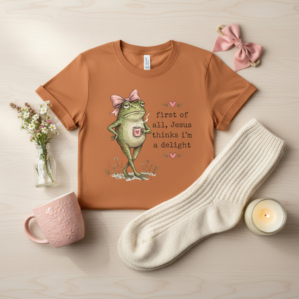 An orange short-sleeve crewneck t-shirt featuring a graphic of a green frog wearing a pink bow and a pink heart-shaped pocket, next to the text 'first of all, Jesus thinks i'm a delight' in black script with small pink hearts.