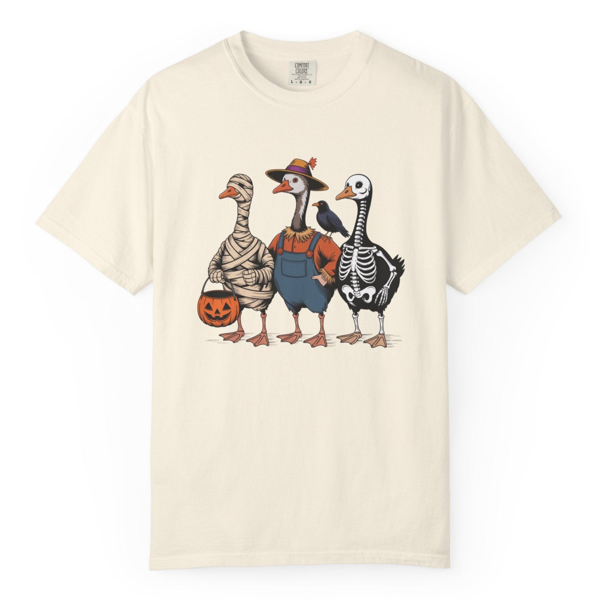 A ivory short-sleeve crewneck t-shirt featuring a graphic print of three cartoonish ducks dressed in Halloween costumes (mummy, skeleton, and farmer boy) holding a jack-o'-lantern bucket.