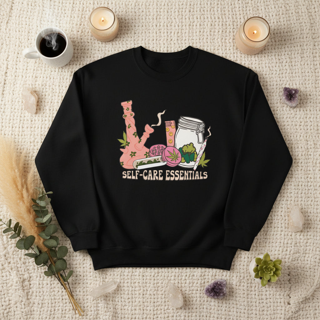 A black crewneck sweatshirt featuring a colorful graphic of self-care and healing items, including a pink bong, a jar, a cup, and a rolling machine, with the text 'SELF-CARE ESSENTIALS' printed below.