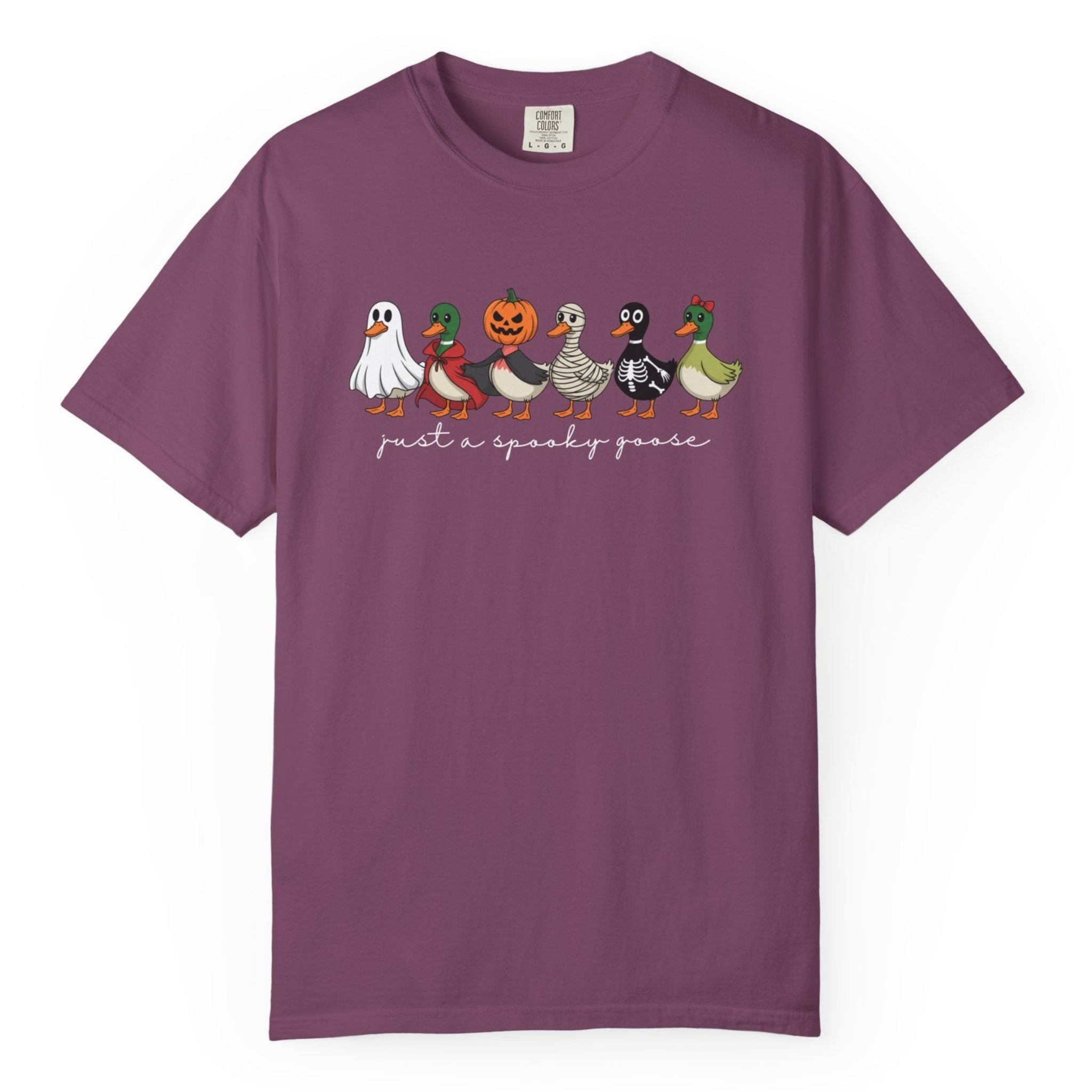 A purple short-sleeve crewneck t-shirt featuring a graphic of six cute cartoon geese dressed in Halloween costumes with the text 'just a spooky goose' printed below them, displayed on a clean white background. 