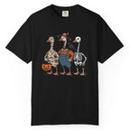 A black short-sleeve crewneck t-shirt featuring a colorful graphic of three geese dressed in Halloween costumes (as a mummy, a farmer, and a skeleton) standing together and holding a jack-o'-lantern.