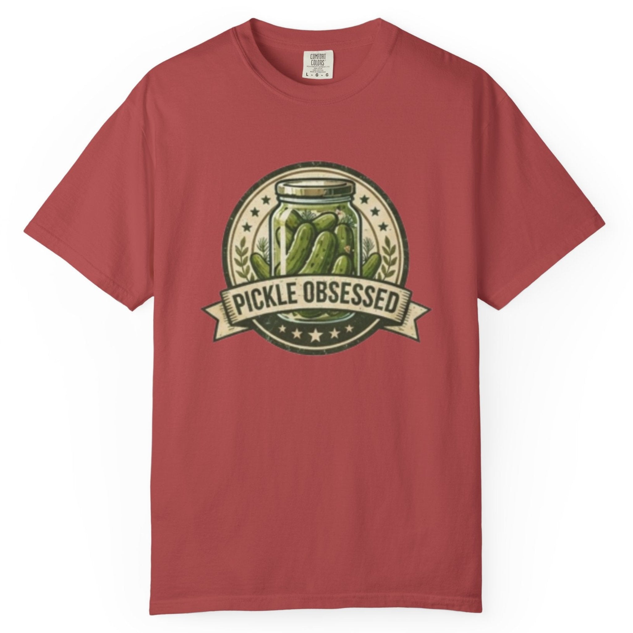 A crimson short-sleeve t-shirt hanging on a wooden hanger against a light wood pegboard wall. The shirt features a circular graphic print with a jar of pickled green onions and the words 'PICKLE OBSESSED' in a banner.