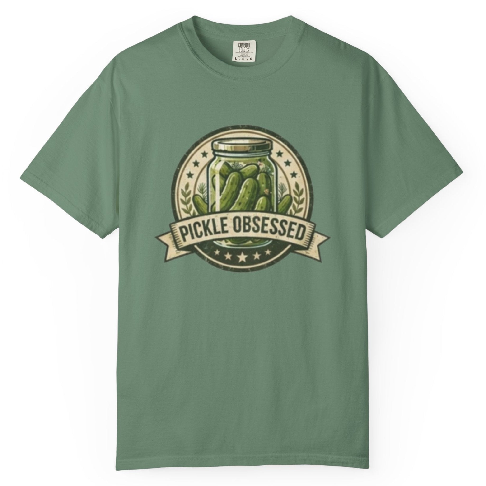 A light Green e short-sleeve t-shirt hanging on a wooden hanger against a light wood pegboard wall. The shirt features a circular graphic print with a jar of pickled green onions and the words 'PICKLE OBSESSED' in a banner.