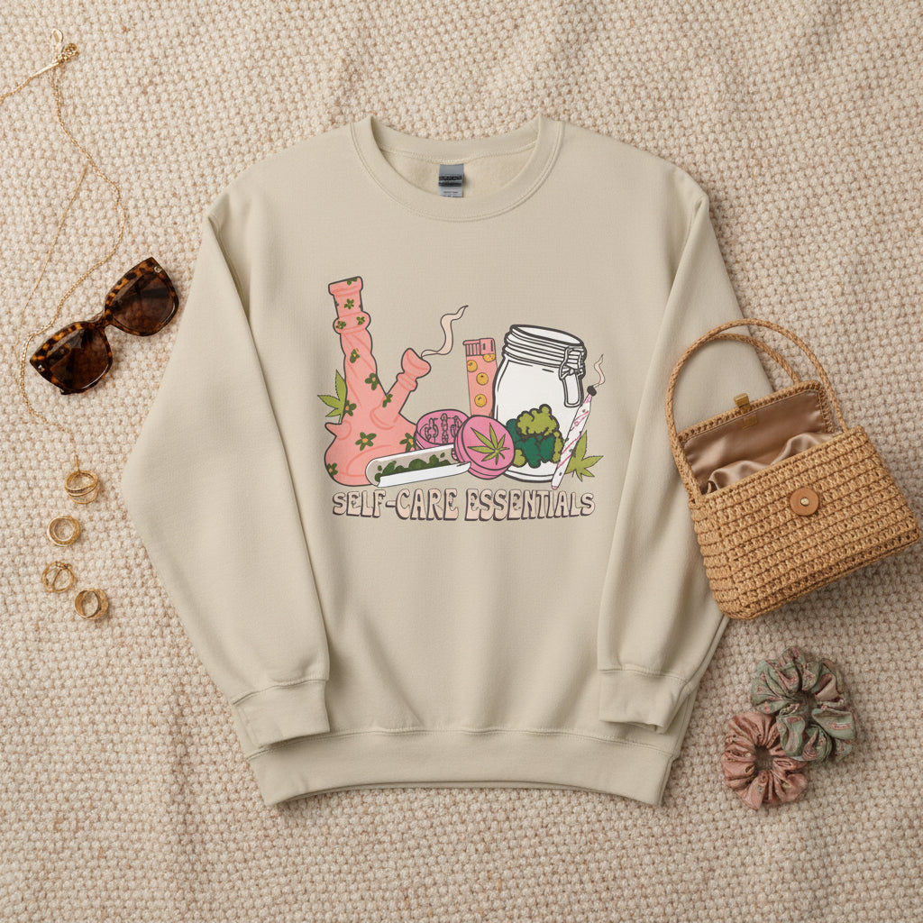 A beige crewneck sweatshirt featuring a colorful graphic illustration of a pink bong, a glass jar, a pink drink, and a green leaf, with the text 'SELF-CARE ESSENTIALS' printed below.