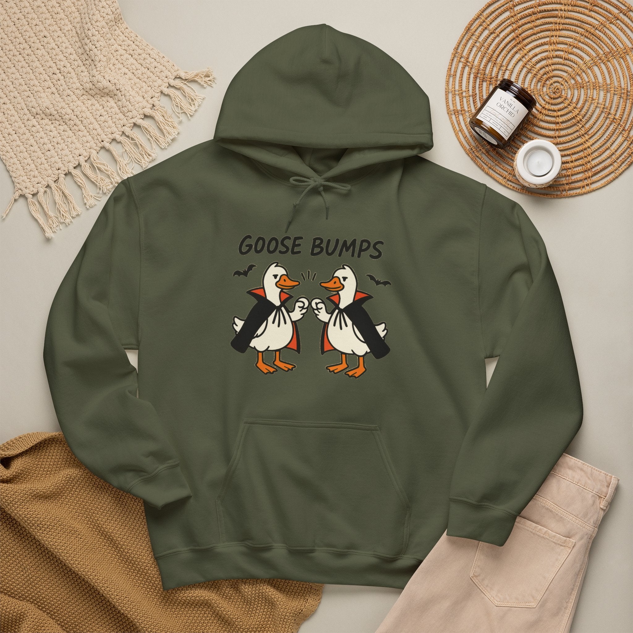 A military green hoodie with a graphic of two geese in black and white costumes with orange beaks, under the text 'GOOSE BUMPS'. The hoodie is styled in a flat-lay arrangement with a beige knit blanket, a brown knit sweater, light pink pants, and a small jar on a woven mat.