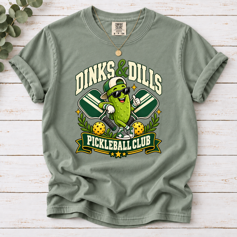 Bay Dinks & Dills Pickleball Club graphic T-shirt featuring a smiling pickle mascot wearing sunglasses and a backward cap, crossed pickleball paddles, and retro collegiate lettering.