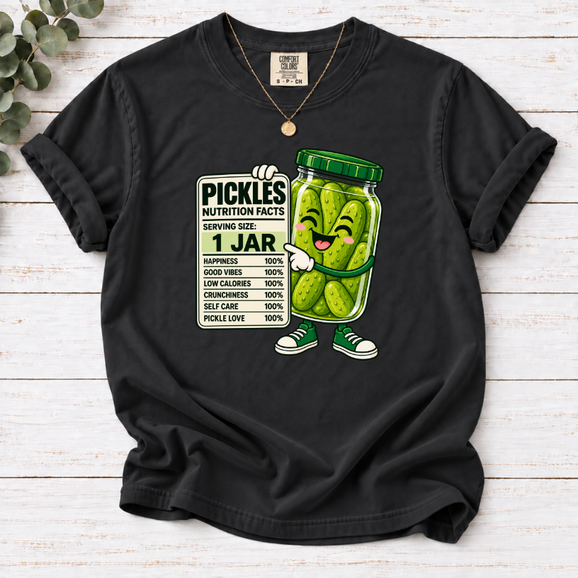A black short-sleeve t-shirt with a graphic of a smiling pickle character next to a nutrition facts label reading '1 JAR PICKLES'. The shirt is displayed flat on a light wood background with eucalyptus leaves in the corner.