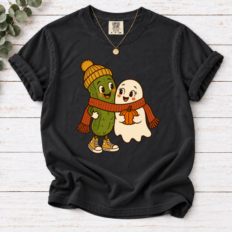 black T-shirt featuring a smiling pickle in a yellow beanie beside a ghost holding a pumpkin, sharing an orange scarf.”