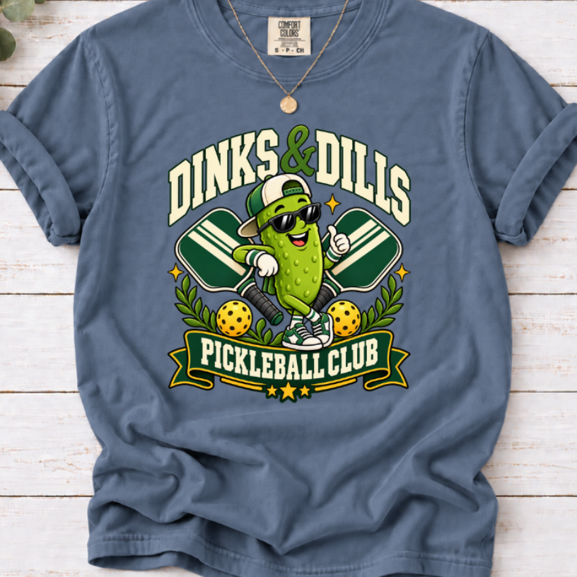 Blue Jean Dinks & Dills Pickleball Club graphic T-shirt featuring a smiling pickle mascot wearing sunglasses and a backward cap, crossed pickleball paddles, and retro collegiate lettering.