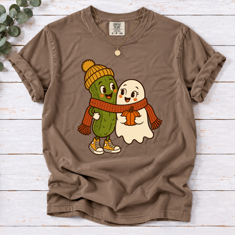 espresso colored T-shirt featuring a smiling pickle in a yellow beanie beside a ghost holding a pumpkin, sharing an orange scarf.”