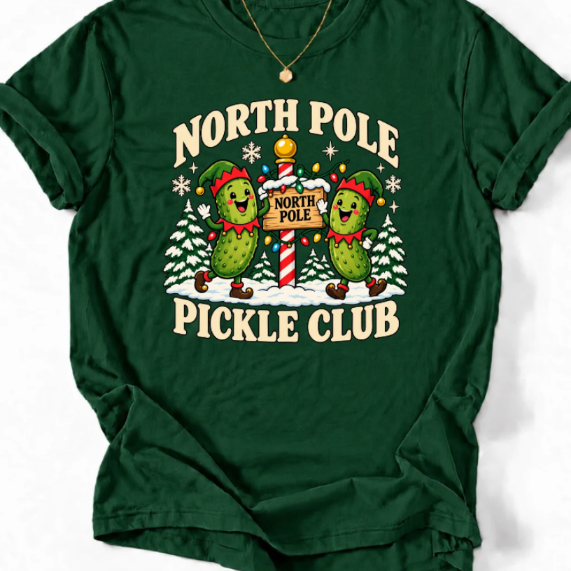 Forest green T-shirt with “North Pole Pickle Club” lettering and two pickle elves beside a candy cane signpost and snowy trees.