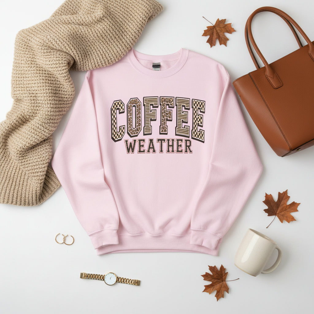 A light pink crewneck sweatshirt featuring the words 'COFFEE WEATHER' printed on the chest in a varsity-style arched font. The letters in 'COFFEE' are filled with a tan and brown leopard print pattern outlined in black, while 'WEATHER' is printed below in solid black.