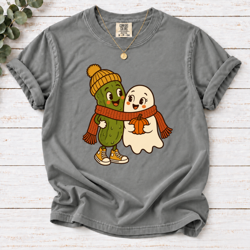 Grey T-shirt featuring a smiling pickle in a yellow beanie beside a ghost holding a pumpkin, sharing an orange scarf.”