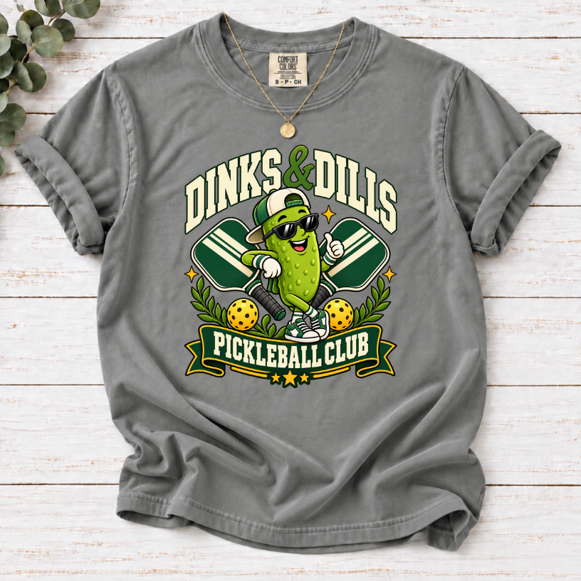 Grey Dinks & Dills Pickleball Club graphic T-shirt featuring a smiling pickle mascot wearing sunglasses and a backward cap, crossed pickleball paddles, and retro collegiate lettering.