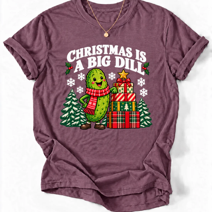 Heather Maroon T-shirt with “Christmas Is a Big Dill” lettering and a smiling pickle in a red scarf beside stacked gifts and snowy trees.