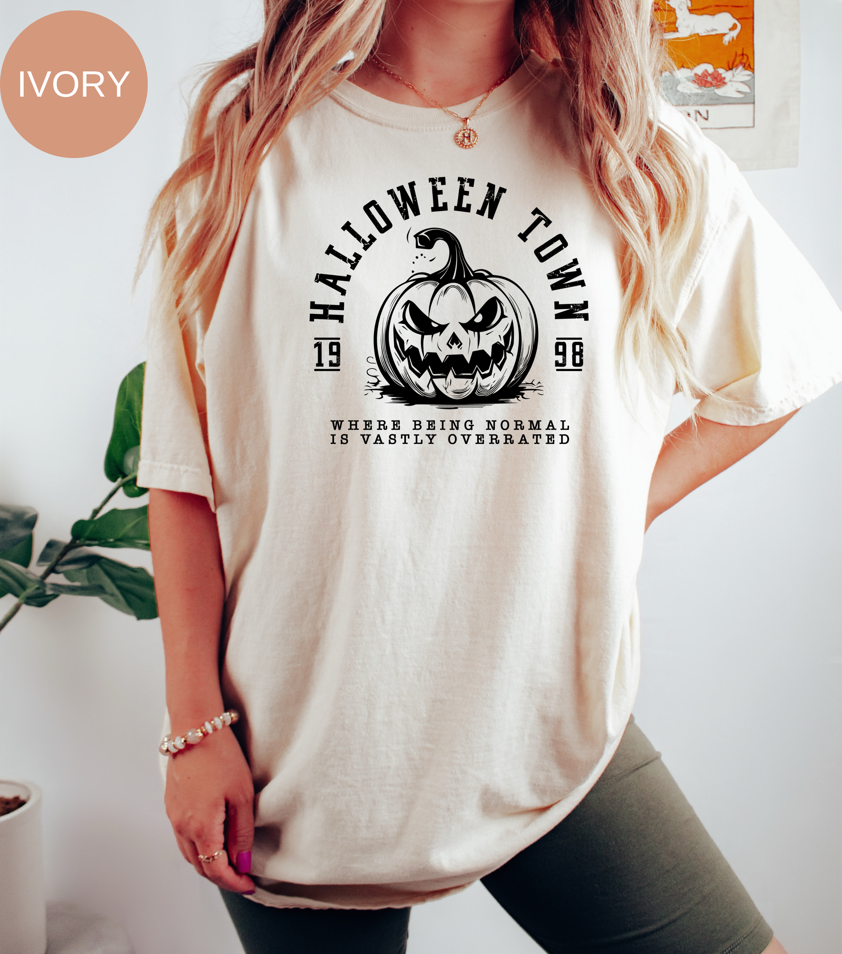 A woman wearing an oversized ivory graphic t-shirt with a black 'Halloween Town 1998' pumpkin design. The image includes an 'IVORY' color label overlay in the top left corner, set against a bright indoor background with a plant.