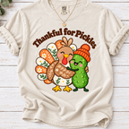 Ivory Thankful for Pickles T-shirt featuring a cartoon turkey hugging a green pickle wearing an orange knit hat.
