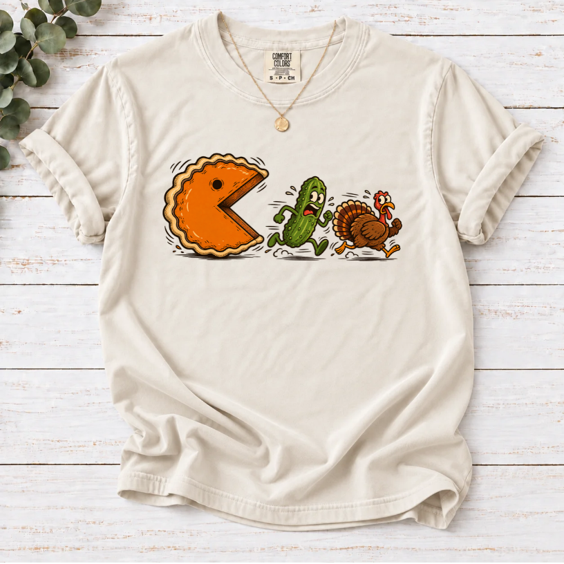 Ivory T-shirt featuring a cartoon pumpkin pie chasing a frightened pickle and turkey.