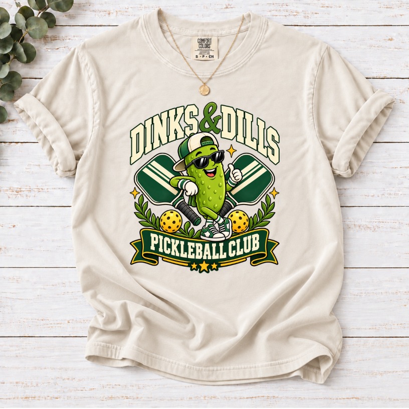 Ivory Dinks & Dills Pickleball Club graphic T-shirt featuring a smiling pickle mascot wearing sunglasses and a backward cap, crossed pickleball paddles, and retro collegiate lettering.