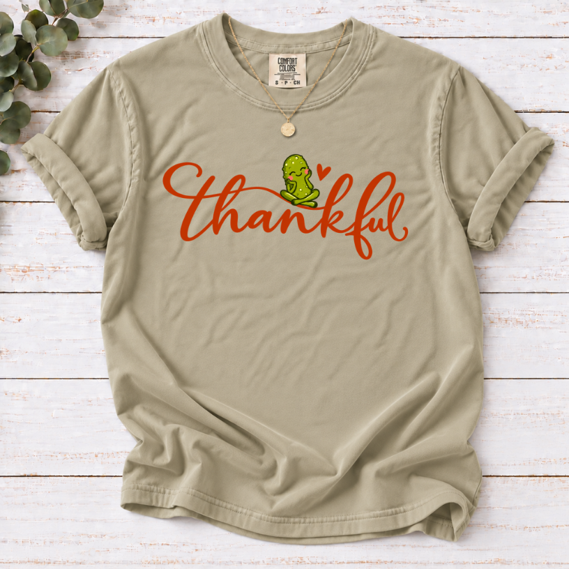Khaki T-shirt with thankful script, a smiling green pickle, and a small orange heart.