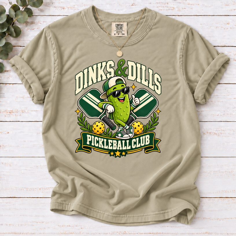 Khaki Dinks & Dills Pickleball Club graphic T-shirt featuring a smiling pickle mascot wearing sunglasses and a backward cap, crossed pickleball paddles, and retro collegiate lettering.